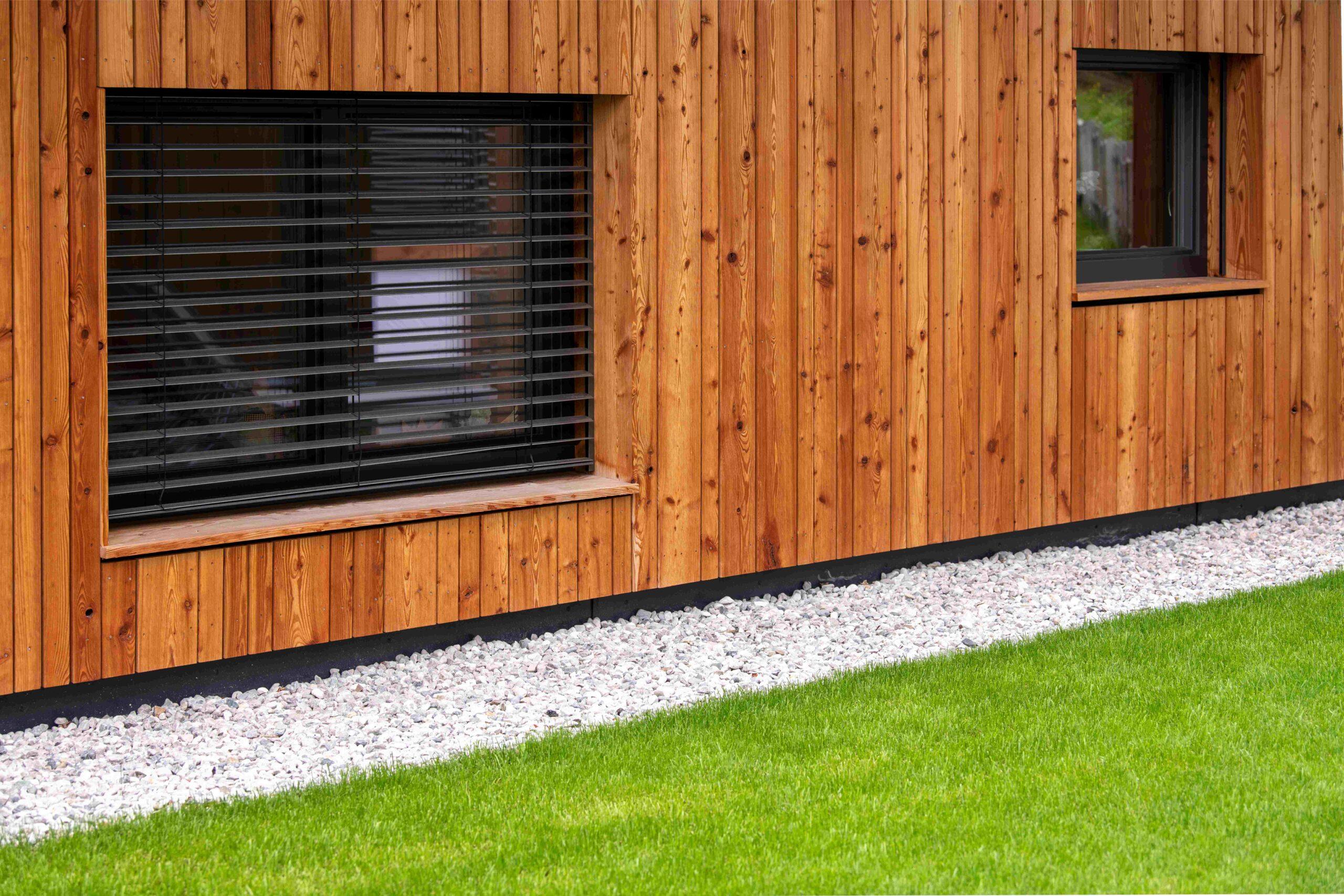 Moderne Architektur mit natürlichen Materialien – Fassade aus Holz, davor sehr gepflegter grüner Rasen und Kiesstreifen – moderne Fenster mit Raffstores 